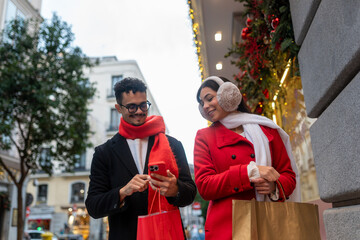 Happy couple engaging with a smartphone while shopping on a festive street in madrid, surrounded by holiday decorations