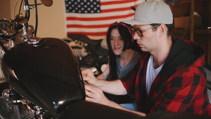 Lovely couple focus intently while repairing a motorcycle in a cozy garage adorned with an American flag, showcasing teamwork and passion for bikes.