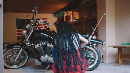 Biker woman with dreadlocks working on motorcycle in garage with American flag backdrop during daytime, showcasing passion for bike maintenance and repair