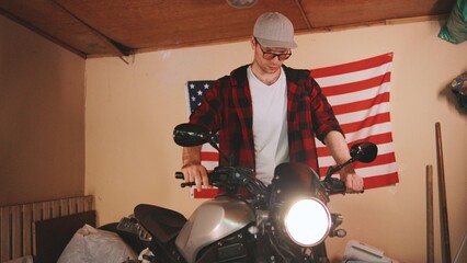 Young man working on motorcycle in garage with American flag in background wearing casual clothes and a cap