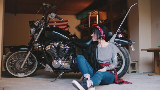 Young woman with long hair sits beside motorcycle in garage, casual outfit accentuated by bandana and tattoos, enjoying a moment of relaxation and reflection - Powered by Adobe