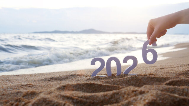 Fototapeta hand place the number 6 on sandy beach, transition from year 2025 to 2026 for new year sign on summer beach at sunset time, with ripple sea wave and beautiful nature at background, New Year celebrate
