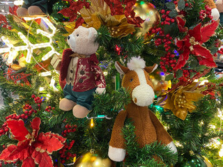 christmas tree with toys. Close-up of a Christmas tree decorated with plush toys &mdash; a bear in a red vest and a brown horse &mdash; surrounded by red poinsettia flowers, gold ornaments, berries, and twinkling
