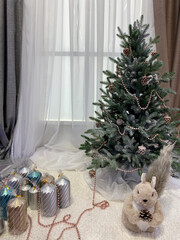 A modern white matte Christmas tree topped with rose gold garland and glittery pine cones sits against a window with sheer white curtains and taupe drapes.