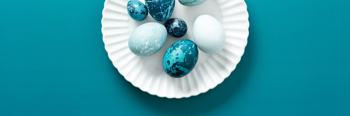 Fototapeta premium Colorful blue Easter eggs are artistically arranged on a white plate resting on a vibrant teal surface, capturing the essence of spring festivities and holiday traditions, banner