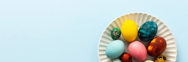 A variety of decorated Easter eggs in vibrant colors are creatively arranged on a white plate set against a soft blue background, symbolizing spring festivities and joy, banner