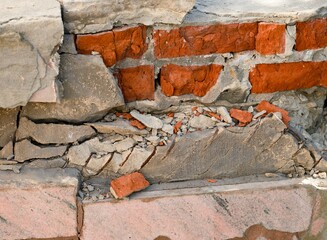 A close-up of a severely damaged brick wall reveals exposed red bricks amidst crumbling, cracked grey mortar and concrete debris.