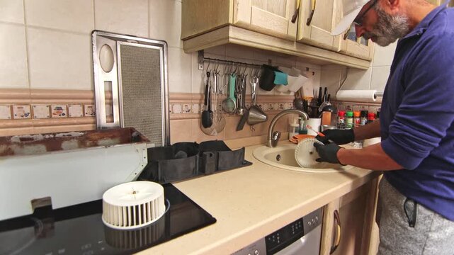 An adult man wearing gloves cleans a greasy kitchen range hood blower fan impeller in the sink, focusing on deep maintenance and hygiene.