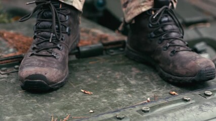 Detailed close-up shot of a soldier's boots and legs in uniform as they step up onto the rugged, metallic armor of a tank or armored fighting vehicle. Symbolizes combat readiness, movement, and milita