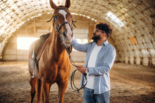 Stroking the animal with his hand. Man is with horse in the hangar - Powered by Adobe