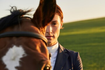 A focused rider, dressed in equestrian attire, looks thoughtfully towards the camera while standing beside a chestnut horse in a grassy field during golden hour.