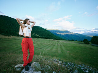 Naklejka premium Outdoor fashion model in a mountain field wearing red pants and white top, hands raised, carefree pose under blue sky, showcasing nature inspired style and rural landscape