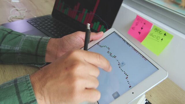 Crypto trader analyzing stock market chart on a digital tablet and taking notes in a notebook