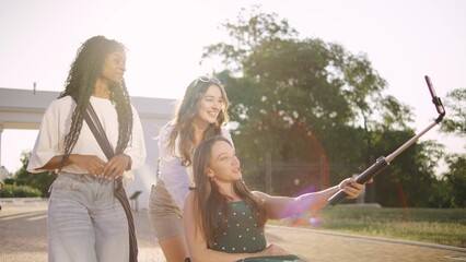 three multiracial female friends one disabled in a wheelchair enjoying a sunny day outdoors while capturing memories with a selfie stick in a park setting