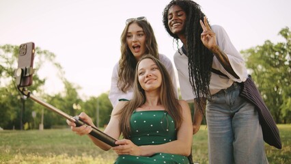 three multiracial female friends one disabled in a wheelchair are enjoying a sunny day in the park taking selfies with a selfie stick while sharing laughter and good times outdoors