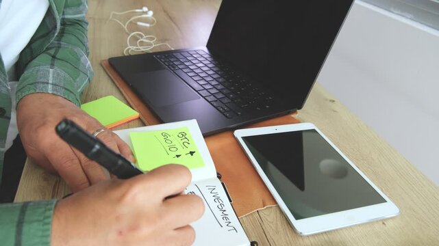 Trader writing investment notes while analyzing financial charts on a laptop and tablet