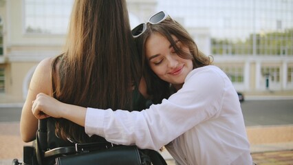two female multiracial friends one inclusive woman in a wheelchair enjoy a sunny day outdoors, sharing hugging and laughter and connection in a joyful moment by a city park