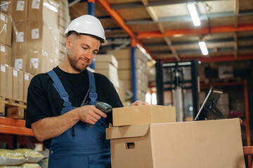 In white hard hat, scanning the box. Man in uniform is working in the storage with packages for shipment