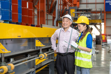 Asian woman worker with helmet and safety vest holds clipboard, discuss with supervisor at industrial site. Senior foreman gives instructions to staff while inspects machinery at manufacturing factory