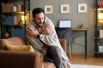 Sick Caucasian man wrapped in a blanket shivering on a sofa at home