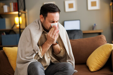 Sick Caucasian man wrapped in a blanket blows his nose at home