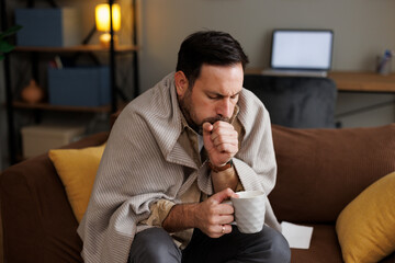 Sick Caucasian man wrapped in a blanket coughs on a sofa at home