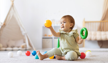 Smiling baby sitting on floor holding colorful ball and ring toy. The bright home interior and...