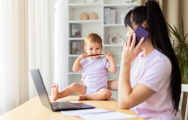 Mother working at laptop while her baby sitting on table chewing pencil. The warm home scene shows multitasking, playful distraction, and modern parenting in a bright lifestyle setting