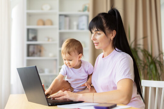 Mother typing on laptop while her baby sitting beside her touching the keyboard. The bright home environment captures multitasking, bonding, and modern family life with warm interaction - Powered by Adobe