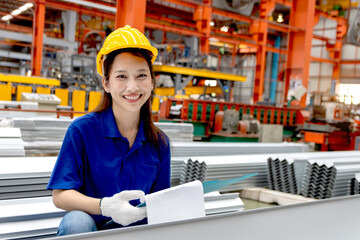 Portrait of happy female Asian worker with helmet holding a clipboard, working at manufacturing factory. Woman staff inspecting metal sheets in industrial site. Inspector checking quality of product.