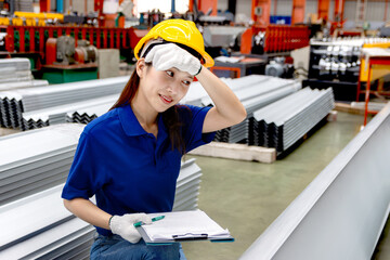 Female Asian worker with helmet and clipboard wiping sweat off forehead, working at manufacturing factory. Tired staff inspecting metal sheets in industrial site. Inspector checking quality of product
