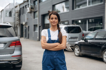 Woman mechanic is standing with arms crossed outdoors against cars and building
