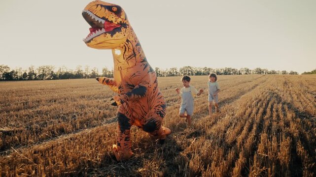 Inflatable Dinosaur T-Rex is Chased, running from Little Kids Boys in field.