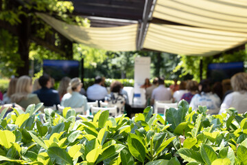 Outdoor business conference with professional audience attending presentation in green garden venue under canopy, sustainable corporate event setting