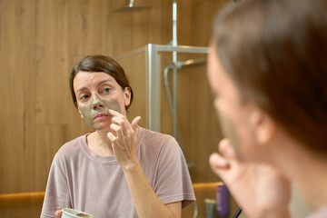 Woman applying clay face mask and looking at reflection in bathroom mirror