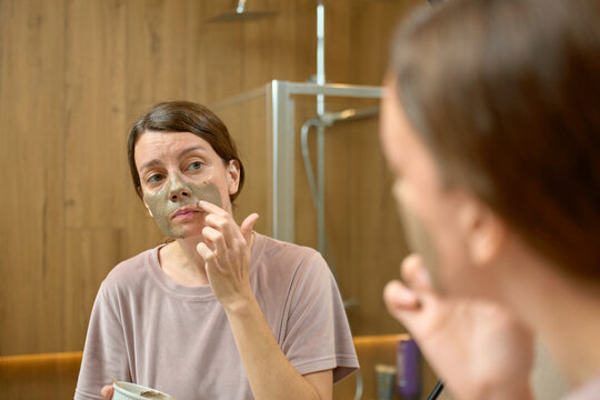 Woman applying clay face mask and looking at reflection in bathroom mirror
