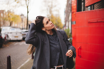Attractive brunette woman walk on the town city street. Girl wear grey wool coat and look happy and smiles. Woman fix hair while go on the street. Girl stand near red door. 
