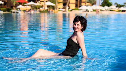 Naklejka premium A brunette woman a black bikini sits in a shallow pool at a luxury resort.
