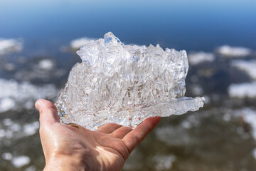A hand is holding a transparent piece of ice, reflecting sunlight, above a calm water surface. The scene captures the beauty of nature and the intricate details of the ice texture and clarity