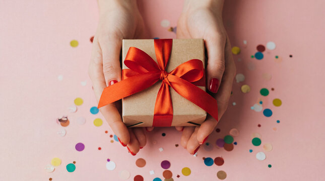 Top view of female hands holding a beige gift box with a red ribbon and bow, surrounded by colorful confetti on a soft pink background. - Powered by Adobe