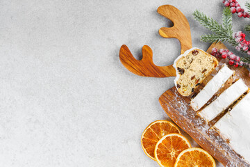 Traditional Christmas stollen, sweet fruit loaf sliced on wooden board, dusted with powdered sugar on white textured background, copy space. Spices, dried fruits and festive decor, winter holiday mood