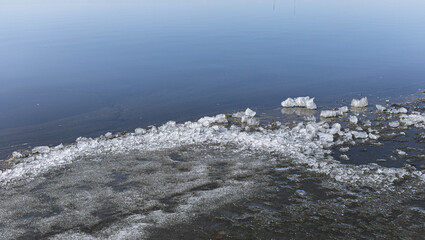 Ice fragments are scattered along the water's edge, creating a serene atmosphere. The calm water reflects the clear blue sky, enhancing the peaceful winter landscape and natural beauty