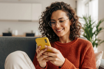 Caucasian woman with curly hair and glasses smiling while looking at yellow smartphone in cozy living room. Digital communication and modern lifestyle, copy space