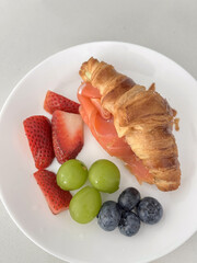 Smoked salmon croissant with fresh strawberries grapes and blueberries on white plate, bright breakfast scene
