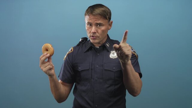 Police officer man points finger to donut while holding it, badge visible on chest, studio setting; duty playful.