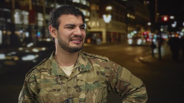 Man wearing camouflage uniform and dogtags grimaces on busy city street at night under glowing streetlights; weariness.