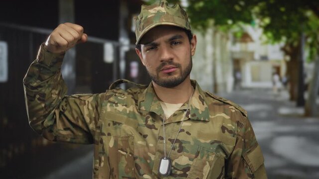 Young hispanic man in camouflage uniform flexes fist showing dog tags on city street; determination.