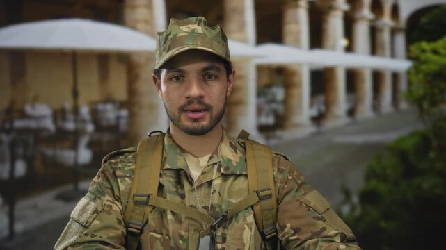 Young hispanic man in camouflage uniform holds hands forward in front of building with visible dogtags; duty honor.