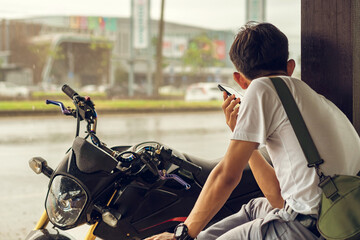 Back view of motorcyclist rests at roadside pavilion and uses smartphone while waiting for rain to stop from heavy rain on side of road. Using communication technology while experiencing travel issues