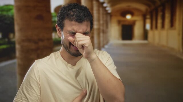 Man clutch chest with one hand and raises other palm toward camera, eyes closed and brows furrowed against pain in old town building walkway; distress.